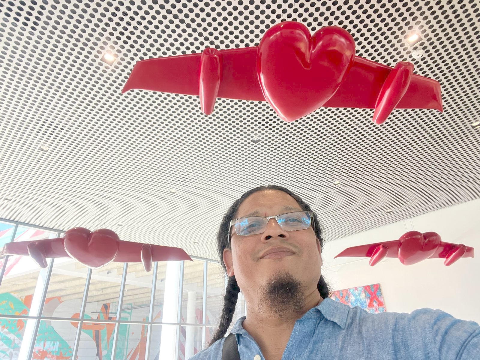 The artist, Esterio Segura, takes a selfie beneath red winged-heart sculptures hanging from a perforated ceiling, with colorful murals visible through the windows behind.