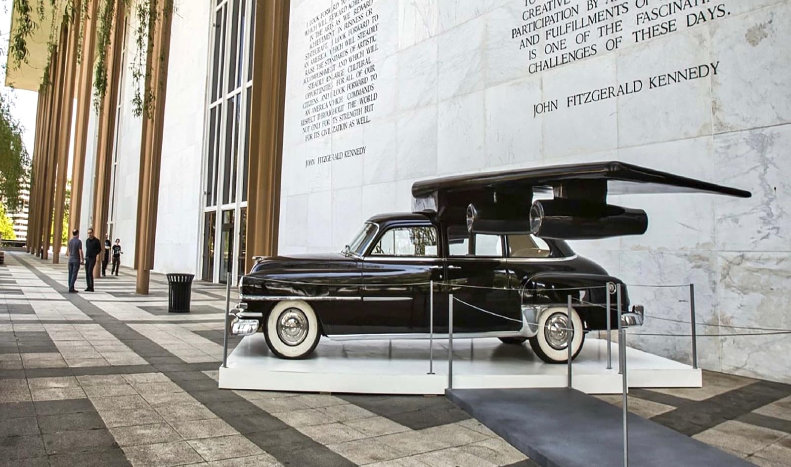 Sculpture of a black vintage sedan with large sweeping wing-like blades rising from its roof, displayed outdoors in front of a colonnade engraved with a John F. Kennedy quote.