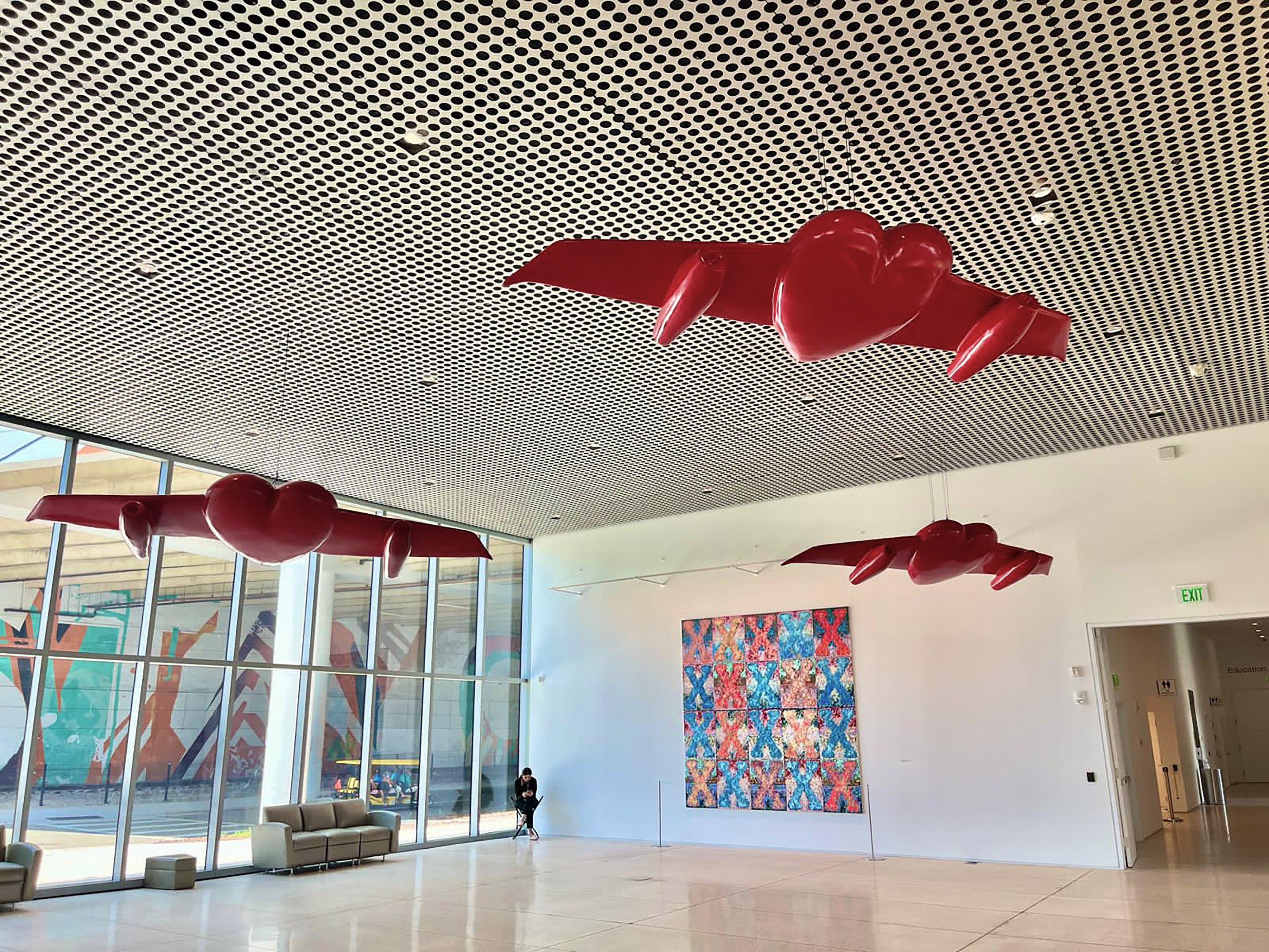 Museum gallery view of three winged-heart sculptures hanging from a perforated ceiling, near a colorful painting and floor-to-ceiling windows.