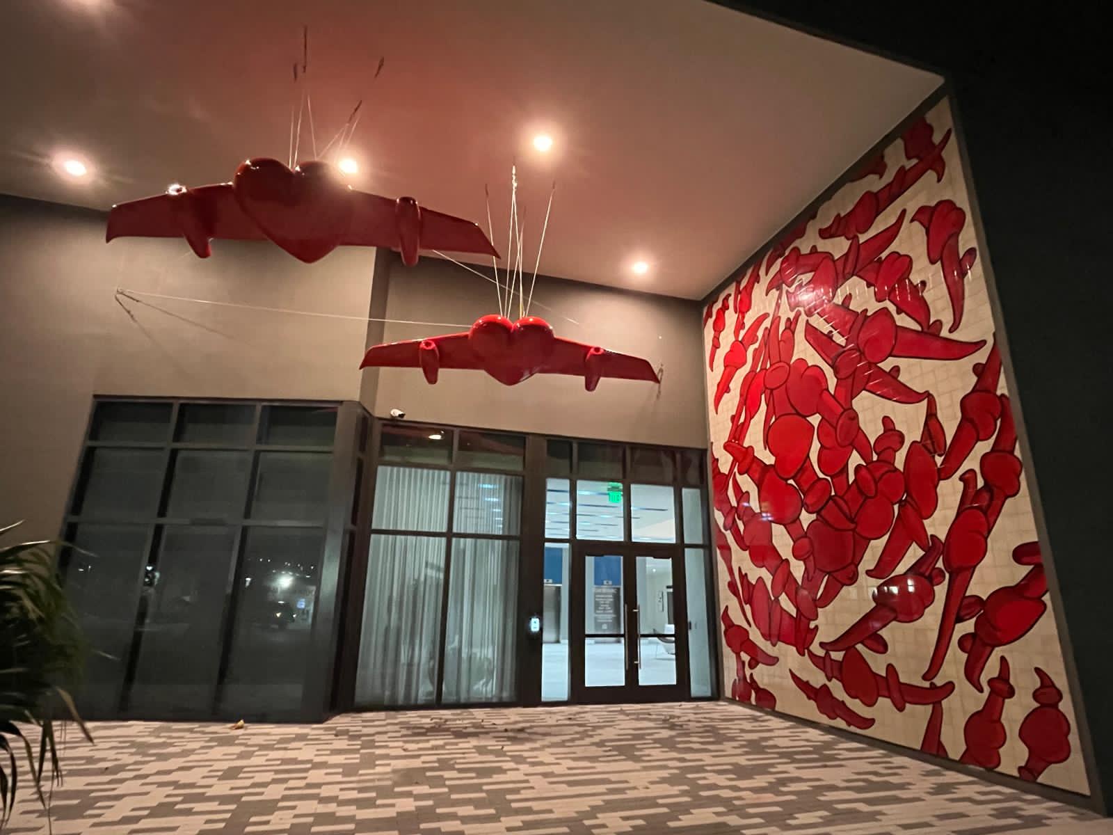 Wide-angle night view of the two suspended winged-heart sculptures above the building entrance, with the red tile mural on the adjacent wall.