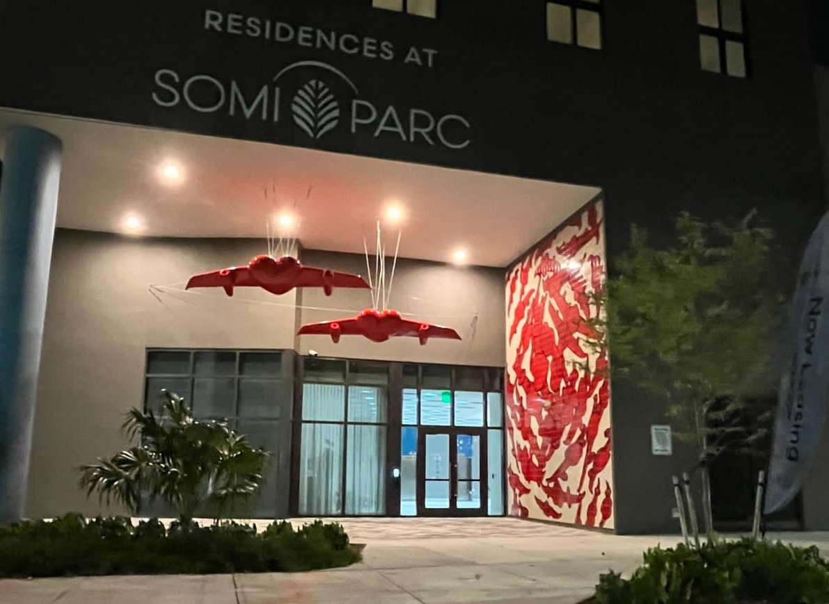 Night view of the SoMi Parc residences entrance, showing the building's signage above the winged-heart sculptures and tile mural.