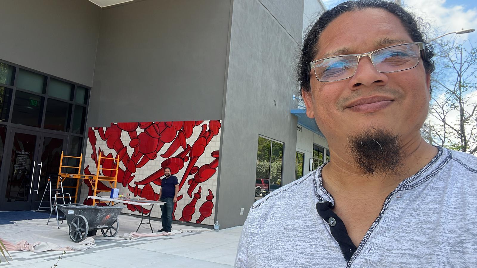 Esterio Segura takes a selfie with the tile mural's installation in progress behind him, scaffolding and tools visible.