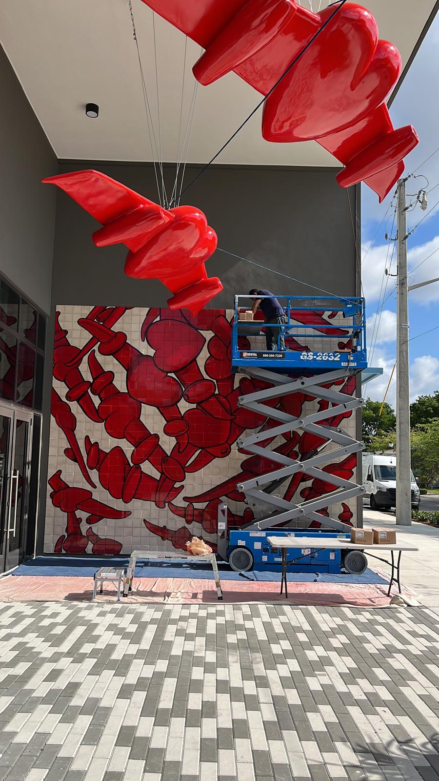A worker on a scissor lift finishes installing the tile mural below the already-hung winged-heart sculptures.