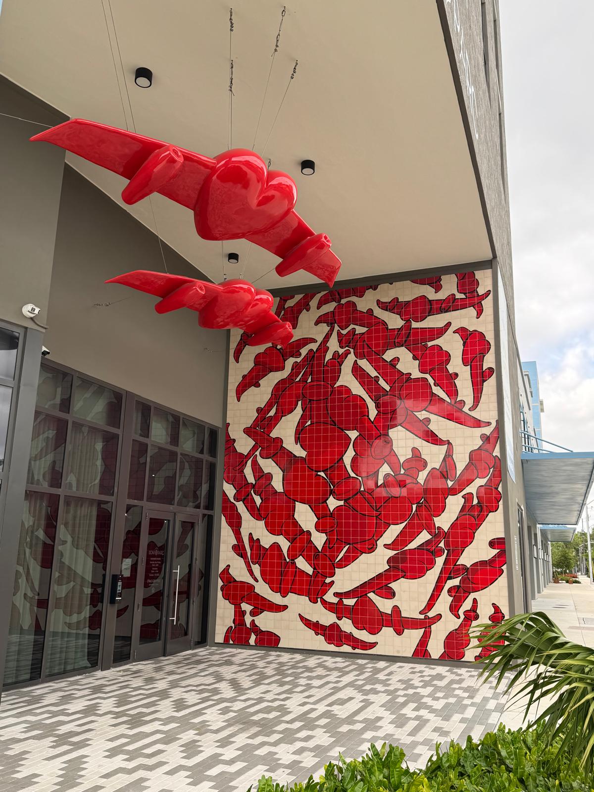 Two red winged heart-shaped sculptures suspended above a building entrance, beside a matching red tile mural of winged hearts on the wall.