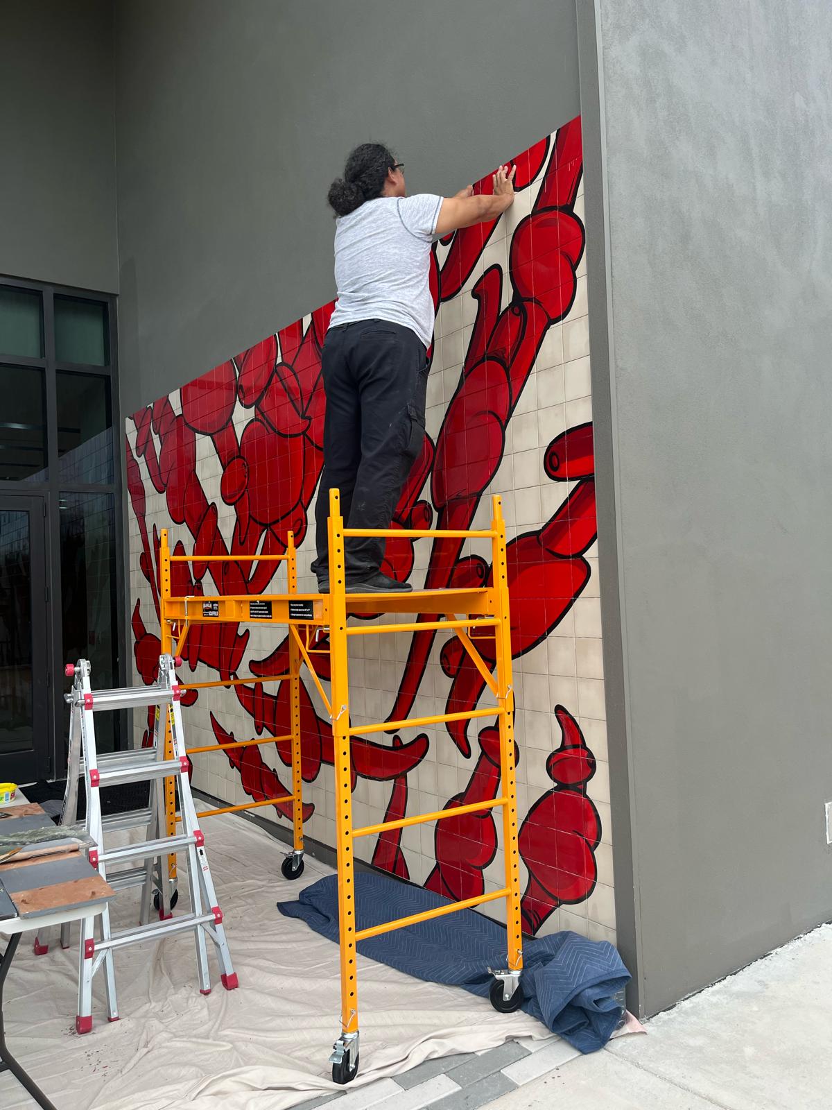 A worker on scaffolding installs the tile mural piece by piece, with the top section still unfinished bare wall.