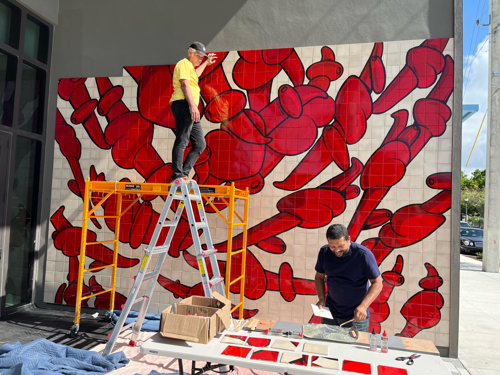 Two workers cut and fit individual red tiles at a table while another attaches them to the nearly complete mural.