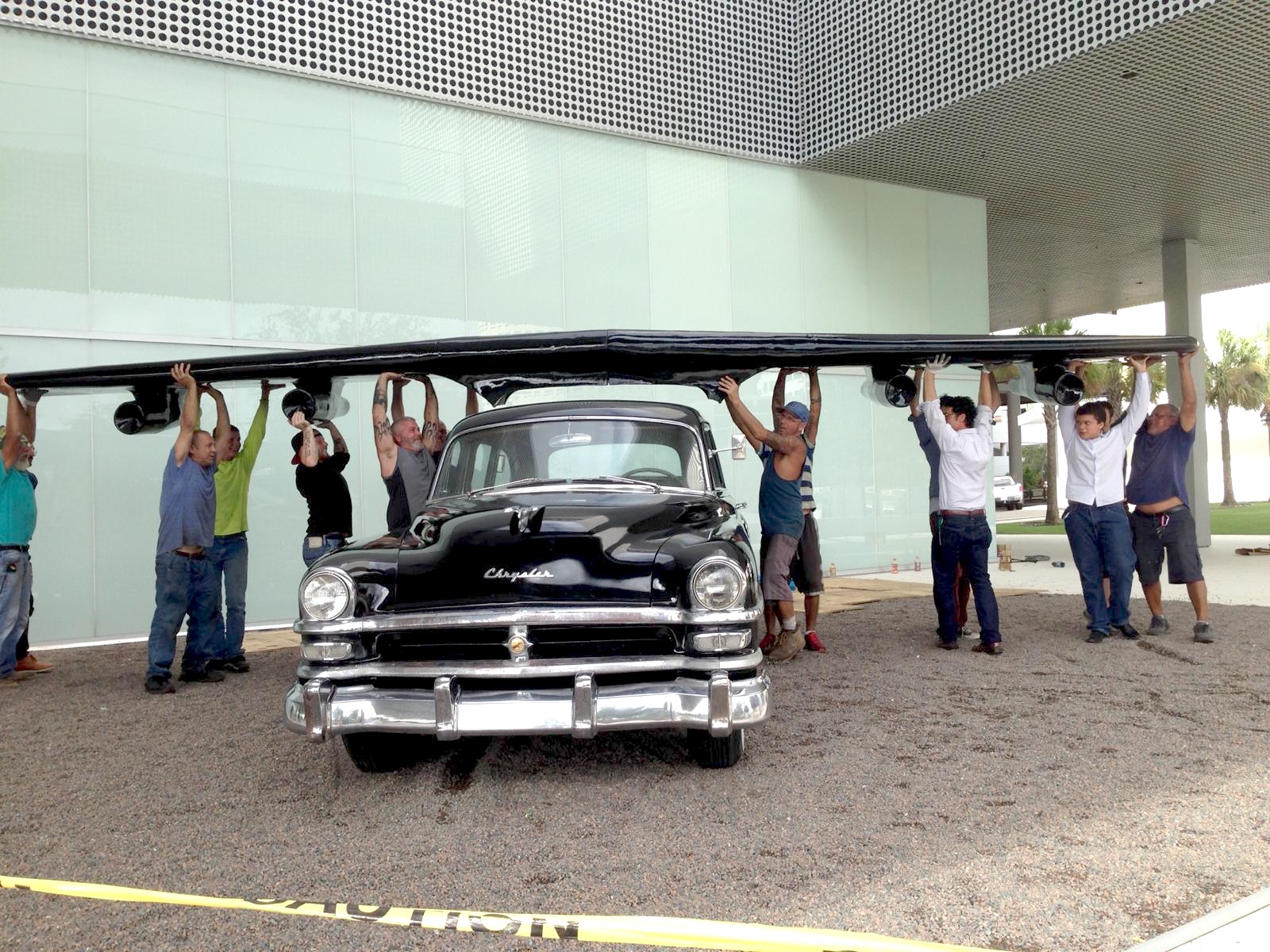 A team of installers lifting the sculpture's large wing section into place above the black Chrysler sedan, outdoors on gravel.