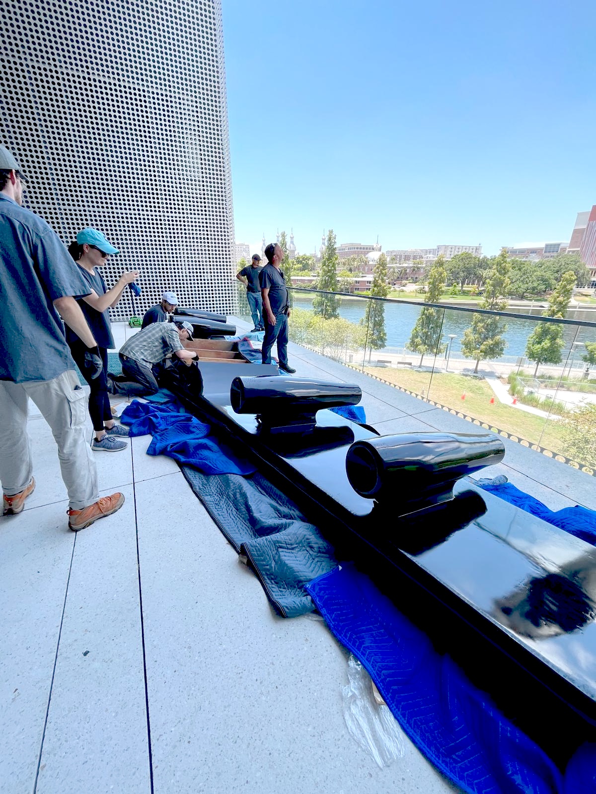The sculpture's black wing section with jet-engine nozzles laid on protective padding during installation, on a terrace overlooking a river.