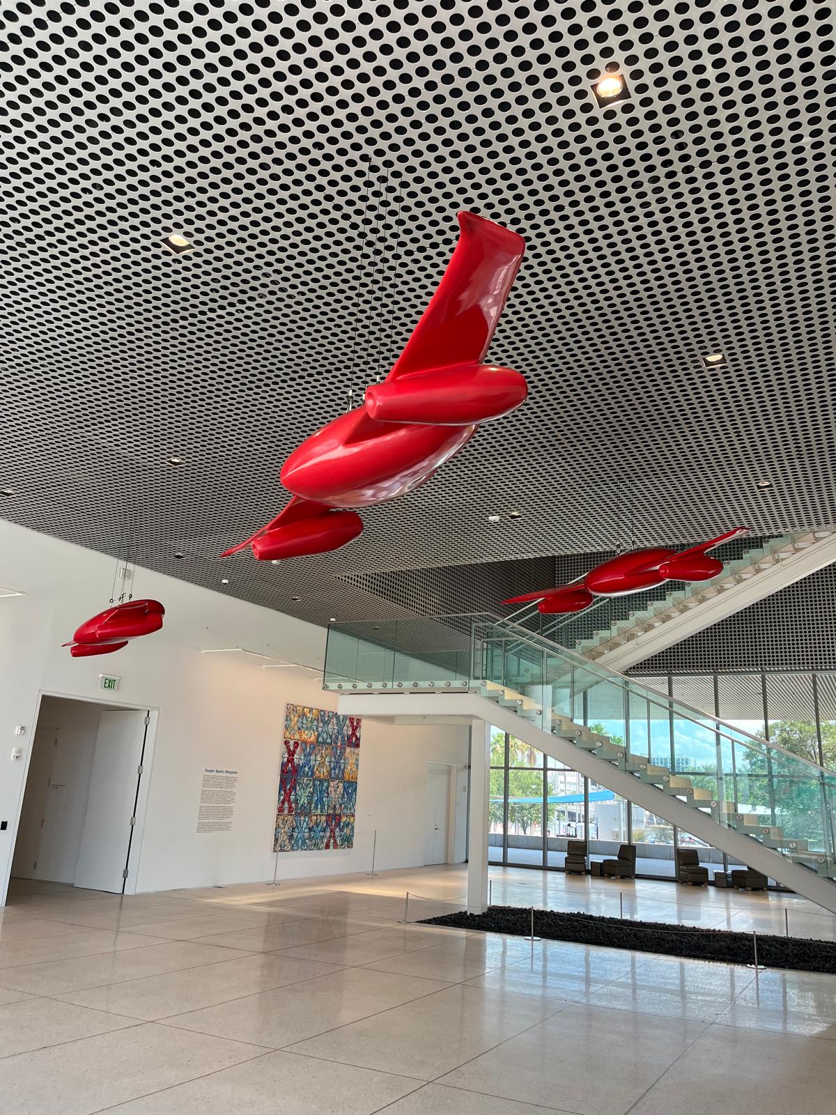 Museum lobby view showing three winged-heart sculptures near the glass staircase and a colorful painting.