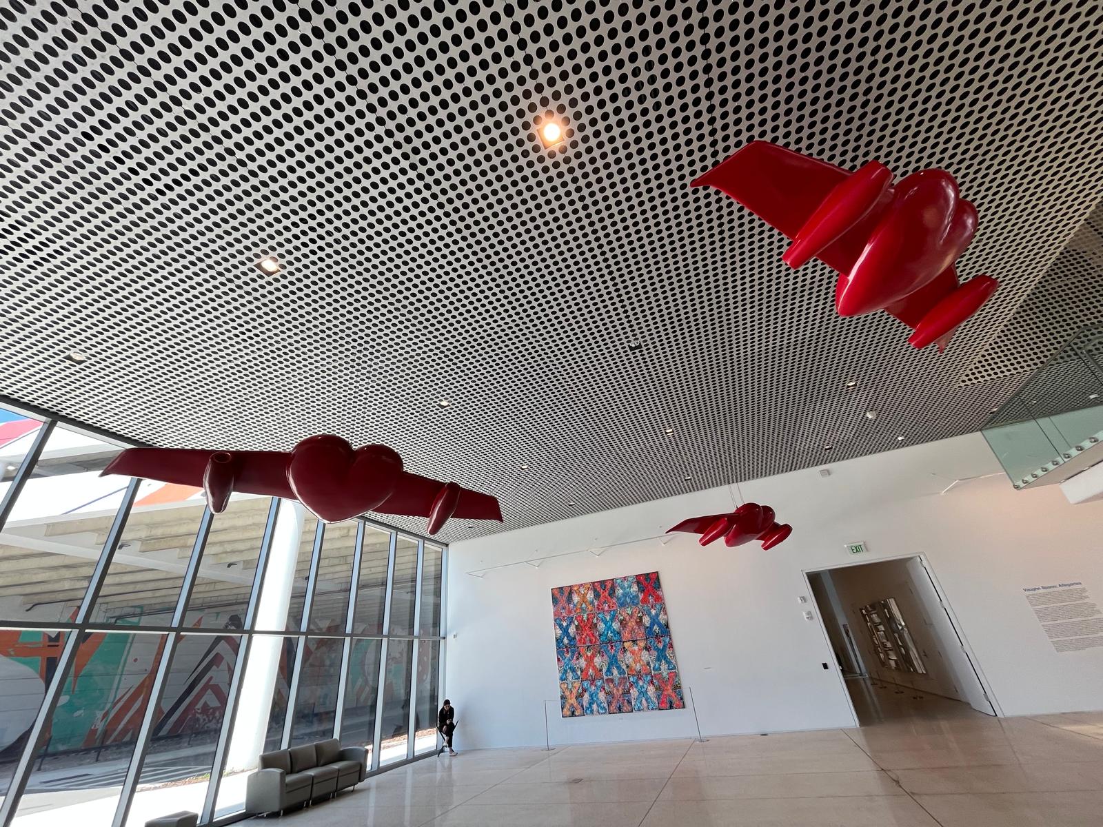 Wide gallery view of three winged-heart sculptures against the perforated ceiling, above the colorful X-patterned painting.