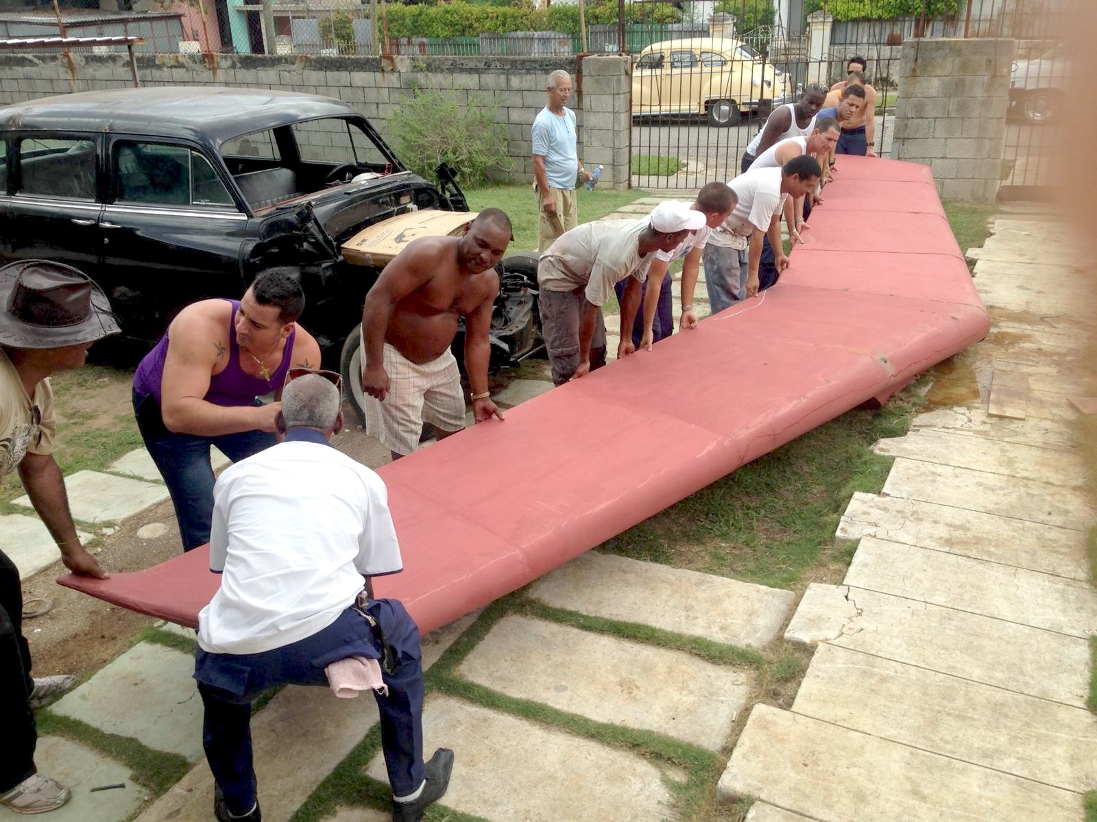 Fabrication studio photo of the sculpture's wing structure in red primer, carried by a group of workers, with a vintage black car nearby.