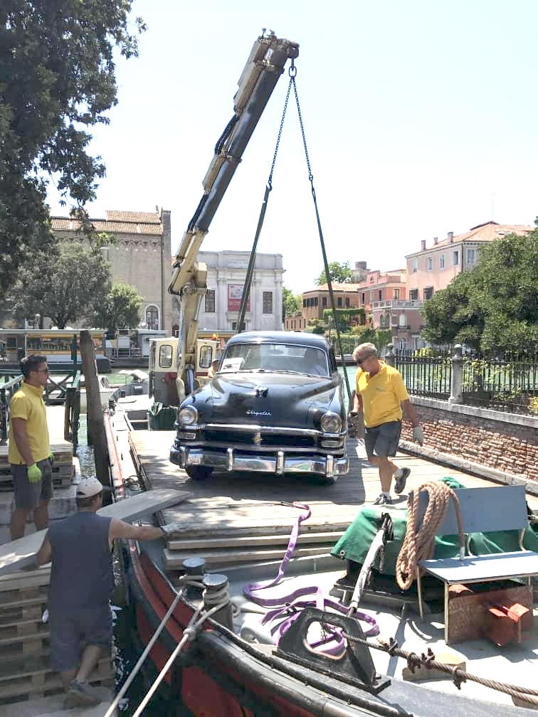 The black Chrysler sculpture being loaded by crane onto a barge along a canal in Venice.