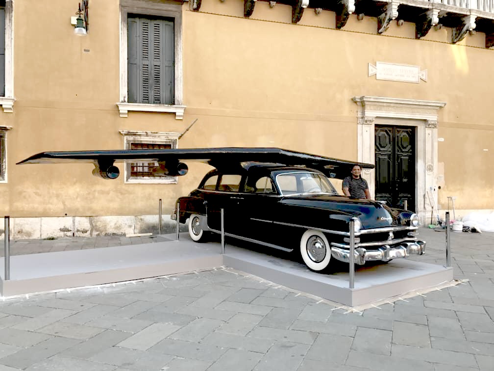 The completed sculpture installed on a roped-off plinth in a Venetian piazza, in front of an ochre palazzo building.