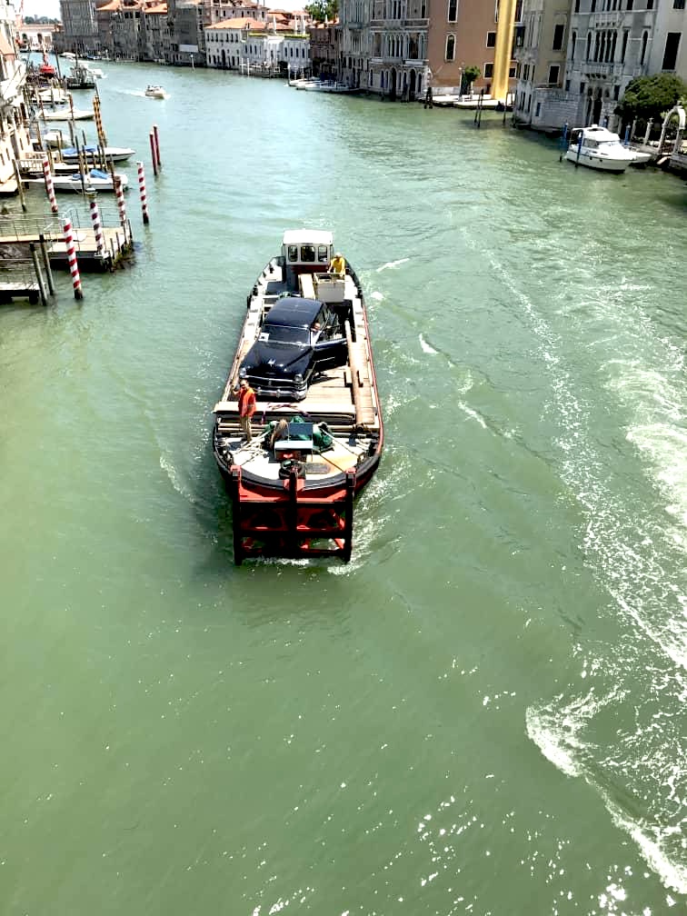 The black Chrysler being transported by barge along a Venice canal, seen from above.