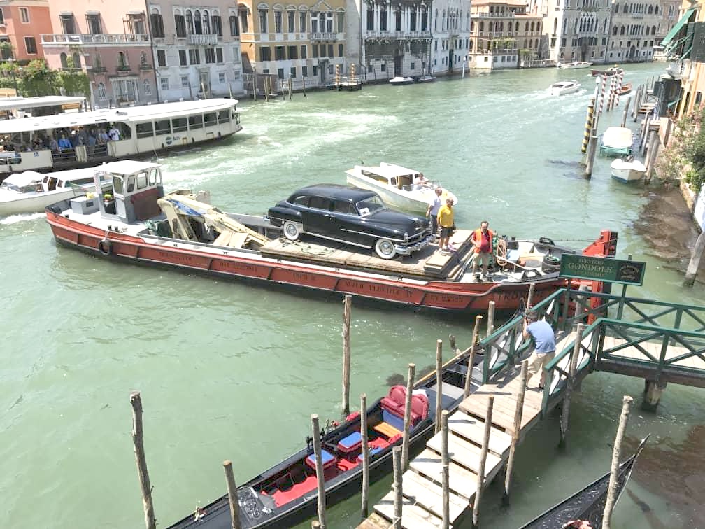 The black Chrysler on a barge navigating the Grand Canal in Venice, with a gondola moored nearby.