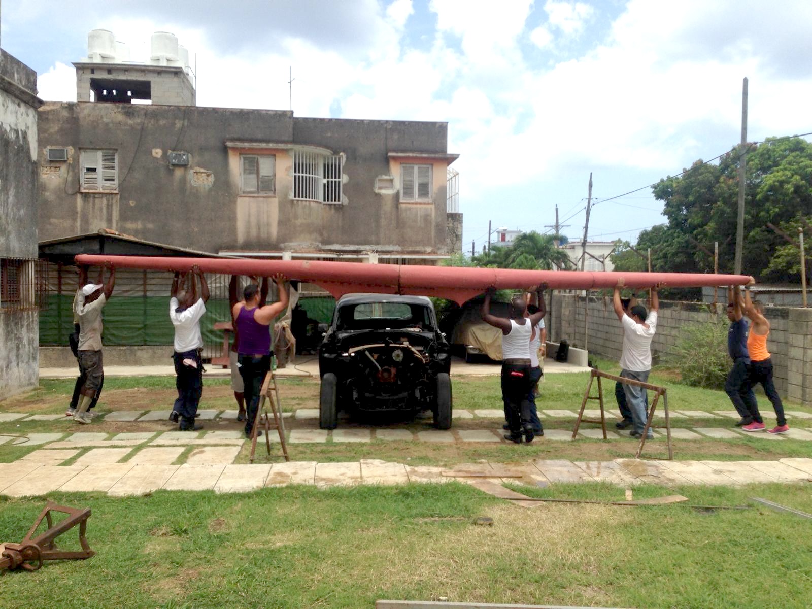 Workers in Cuba lifting the unpainted wing structure over the car's disassembled chassis, in a courtyard with weathered buildings.