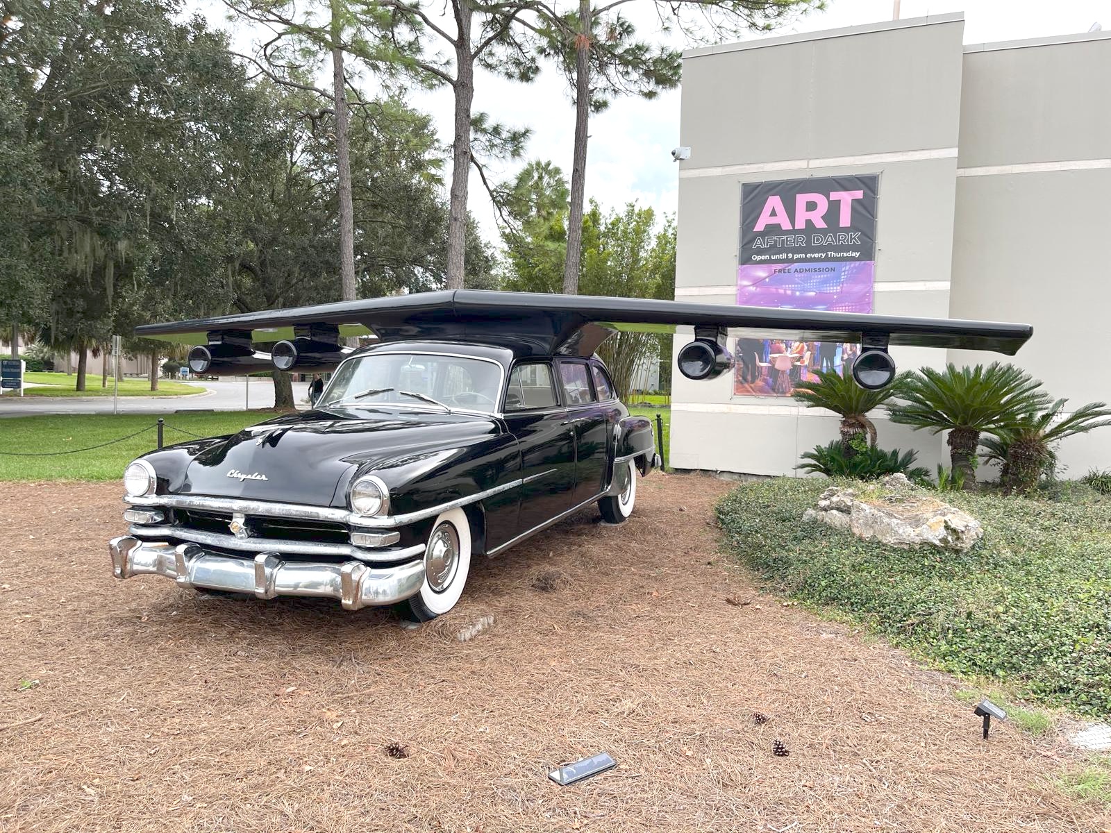 The sculpture on display outside a museum entrance with an 'Art After Dark' banner, surrounded by pine trees and palms.