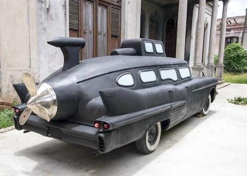 Sculpture combining a vintage American car with submarine features — a propeller, periscope, and round portholes — parked outside a colonial-style building.