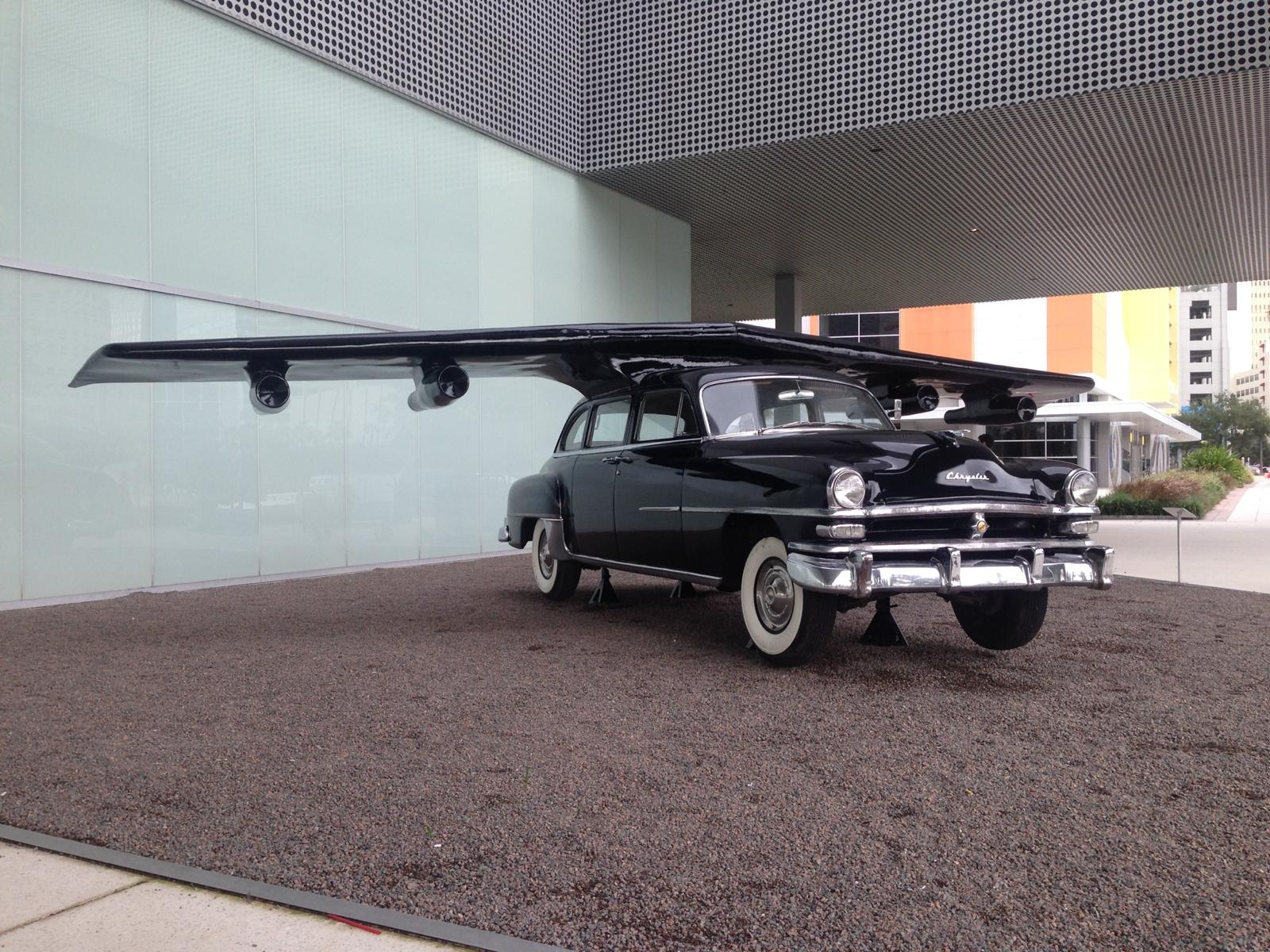 Sculpture of a black vintage sedan with a wide wing bearing jet engines mounted on its roof, displayed outdoors on gravel beside a modern museum building.