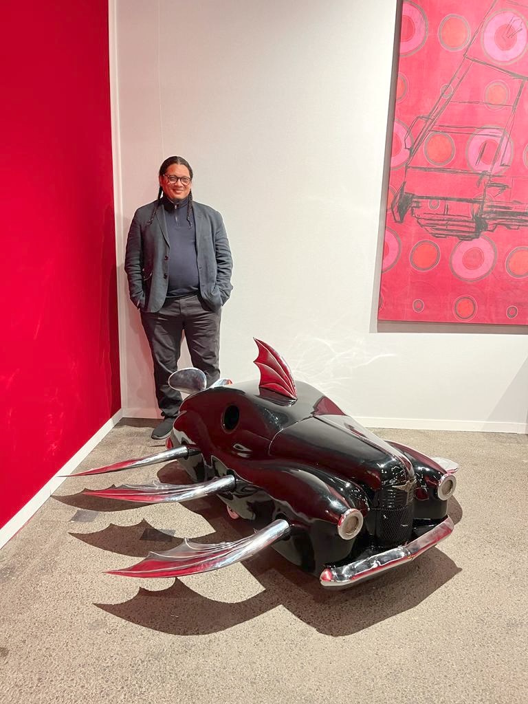 A man stands beside a dark red car-front sculpture with bat-like chrome fins, in a gallery with a red accent wall and a red circular-patterned painting.
