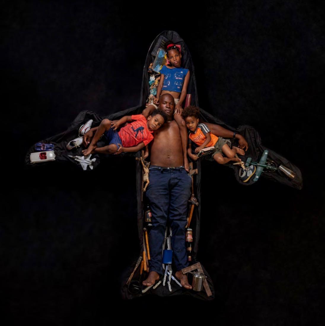 A man and three children posed within an airplane-shaped black suitcase sculpture, surrounded by small personal objects and toys arranged along its wings and body.