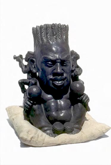Dark bronze sculpture of a stylized figure with a spiky crown-like hairstyle and multiple small arms, resting on a cream-colored cushion.
