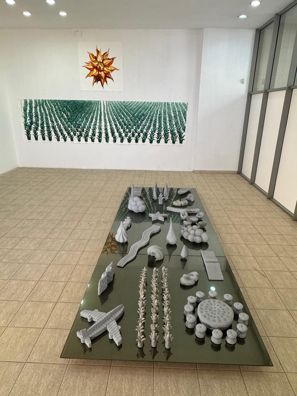 A gallery installation of many small white miniature sculptures arranged on a long glass table, beneath paintings of a starburst and a green field.