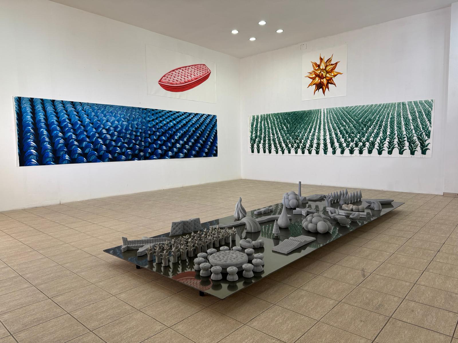 Full gallery view of the same installation, with paintings covering two walls and the miniature sculptures displayed on a long glass table.