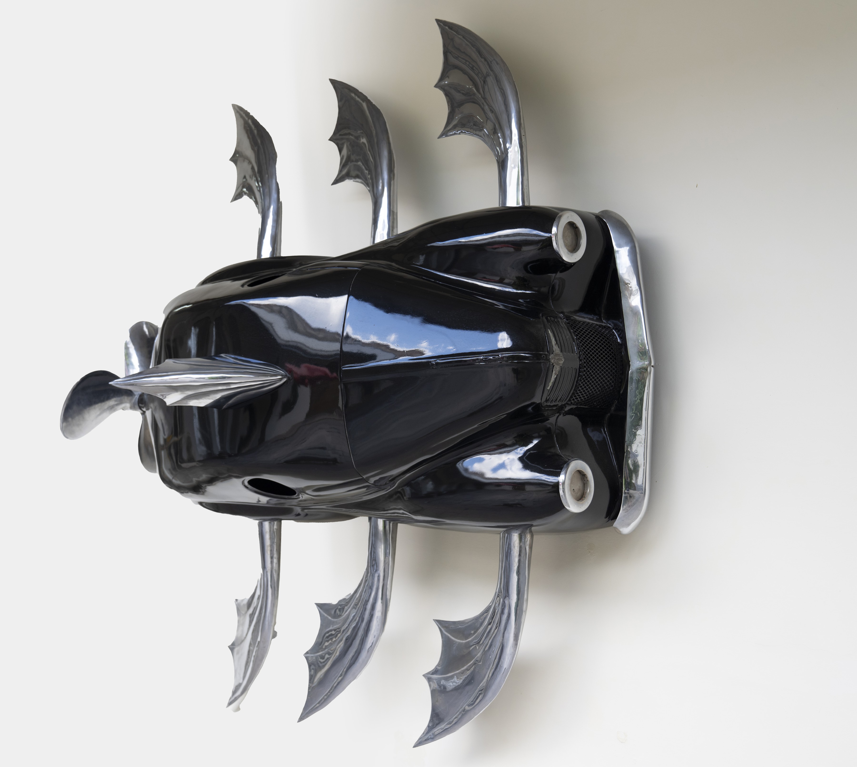 Wall-mounted sculpture resembling a stylized fish assembled from chromed vintage automobile parts, with a glossy black teardrop body, a chrome spear-like nose, round headlight eyes, and curved chrome fin shapes radiating from the top and bottom.