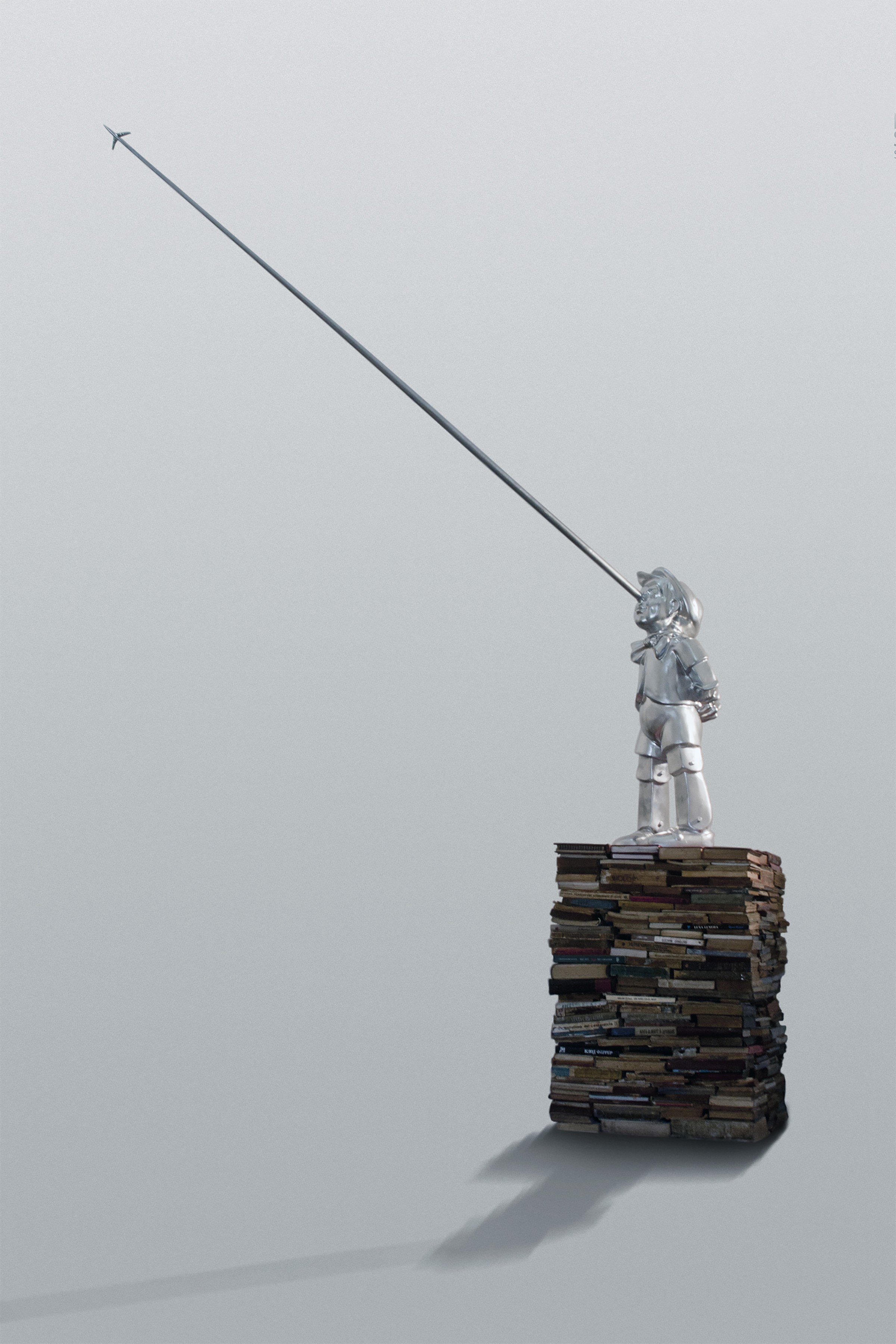A chrome Pinocchio-like figure standing on a stack of books, its elongated nose extending upward to a tiny airplane.