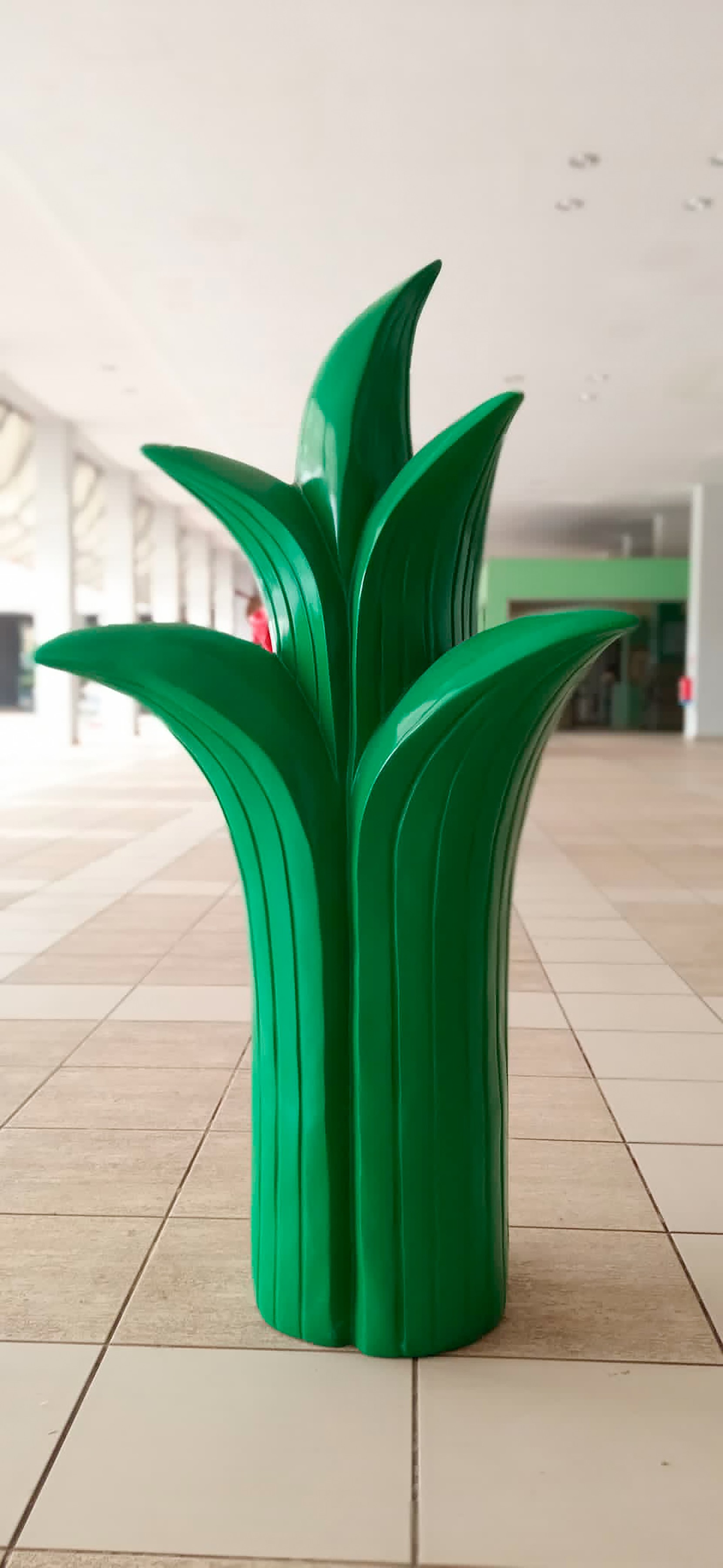A glossy green sculpture shaped like a stylized plant frond, standing on a tiled floor in an indoor arcade.