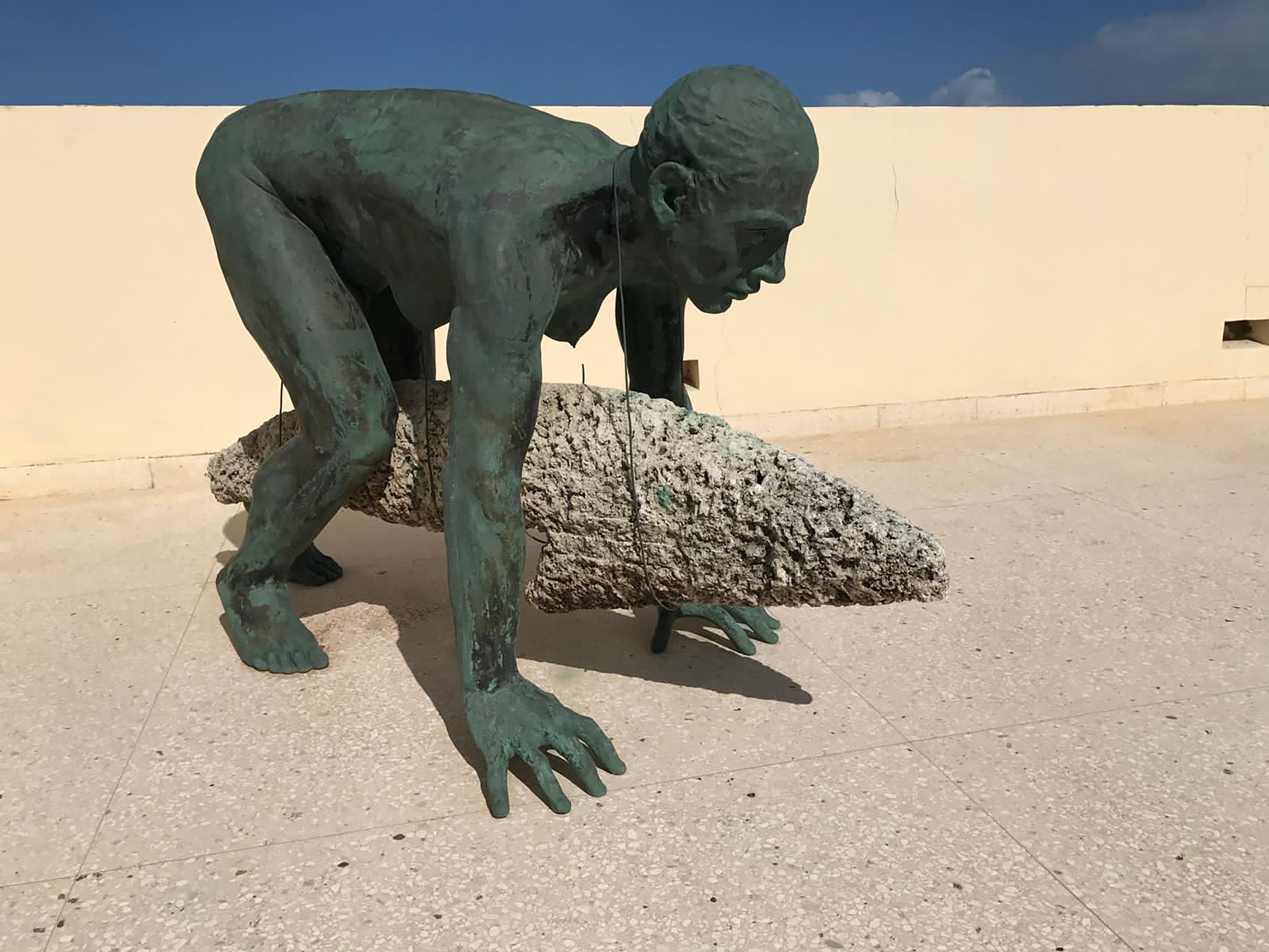 A bronze sculpture of a crouching nude figure whose lower body transitions into a rough stone, animal-like tail, on an outdoor terrace.