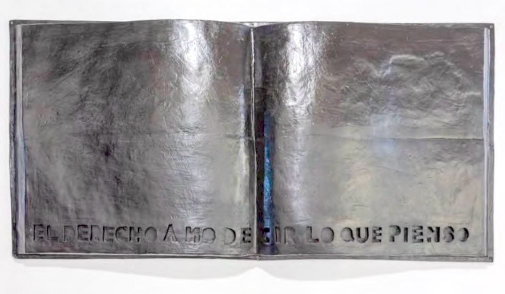 Metal relief sculpture shaped like an open book with hammered, wrinkled silver pages, the Spanish phrase 'EL DERECHO A NO DECIR LO QUE PIENSO' cut into the bottom edge.
