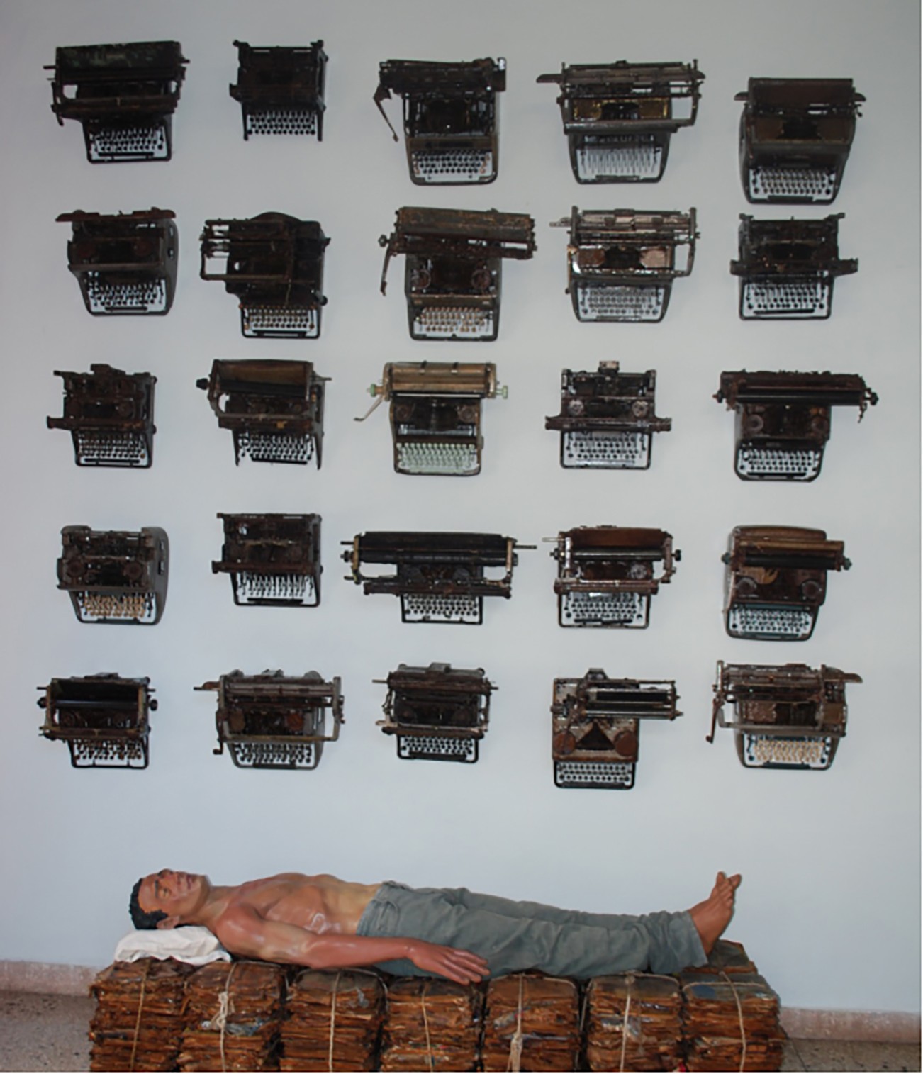 Rows of rusted vintage typewriters mounted on a wall above a man lying on a bound stack of bundles.