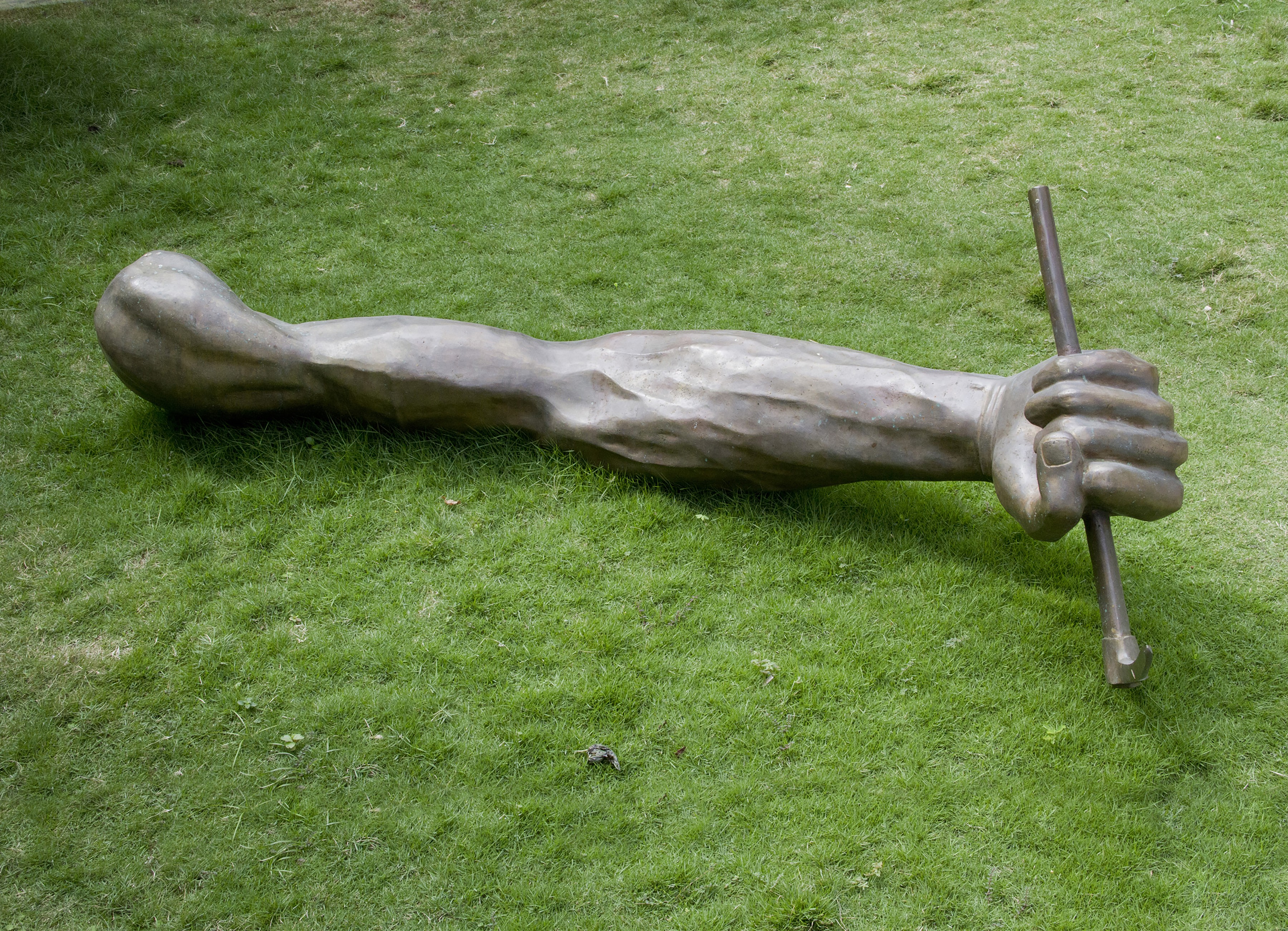 Bronze sculpture of a life-size human arm and clenched fist lying in the grass, gripping a metal rod planted in the ground.