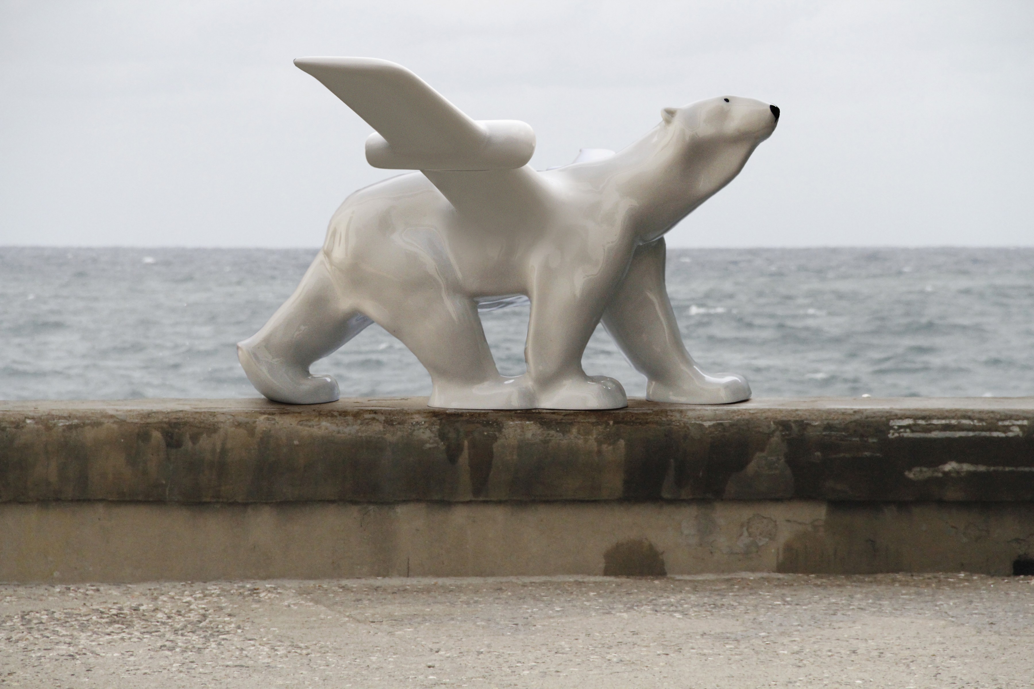 Outdoor sculpture of a white ceramic polar bear walking along a stone ledge by the sea, with a pair of wing-like fins rising from its back.
