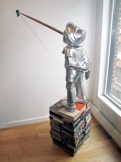 The chrome Pinocchio-like figure on its book pedestal, shown indoors near a window.