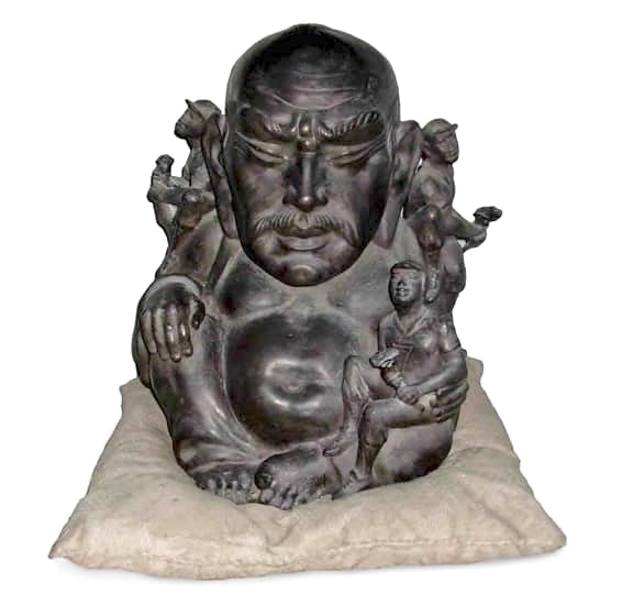 Bronze sculpture of a bald, mustached head and torso seated on a cushion, with several small figures perched around its shoulders and one child figure held against its side.