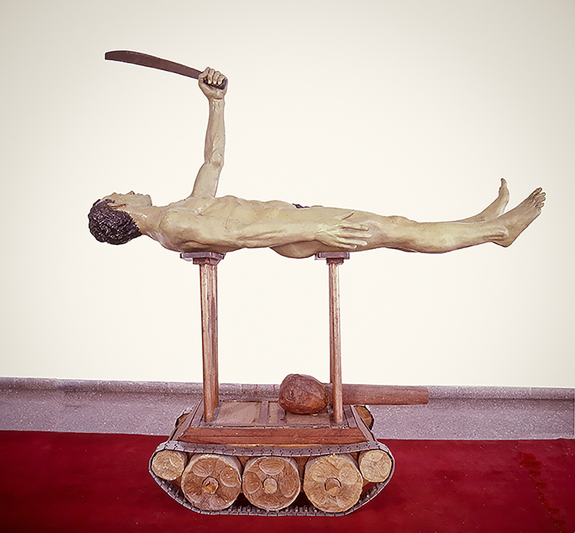 Sculpture of a nude male figure lying flat on his back atop a pair of parallel wooden beams set on a log-rolling base, one arm raised holding a curved blade.
