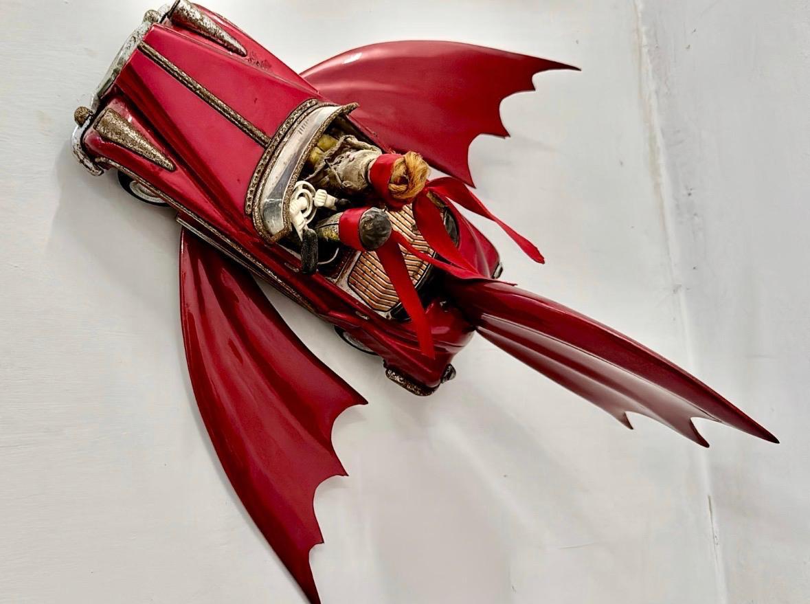Sculpture of a vintage red convertible car fused with bat-like wings and a fish tail, its open cabin cradling wrapped bundles tied with a red ribbon, mounted on a white wall.