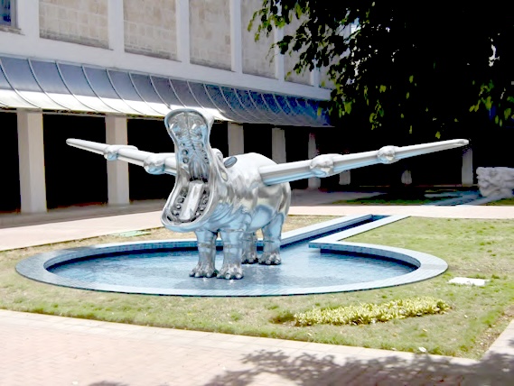 Outdoor sculpture of a silver hippopotamus with open jaws and long wing-like arms extended outward, standing in a shallow reflecting pool outside a building.