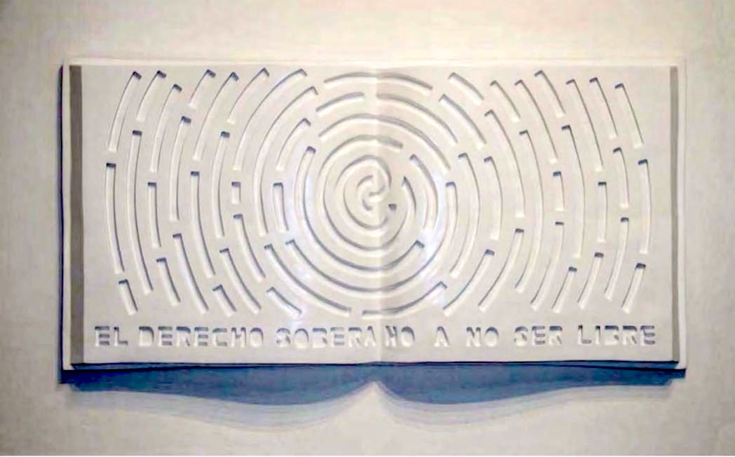 White relief sculpture shaped like an open book, its pages carved with a radiating spiral maze pattern and the Spanish phrase 'EL DERECHO SOBERANO A NO SER LIBRE' along the bottom.