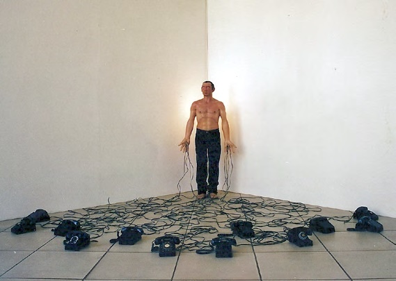 Photograph of a shirtless man standing barefoot in the corner of a white gallery room, dozens of black rotary telephones scattered on the floor around him, their cords trailing to his outstretched hands.