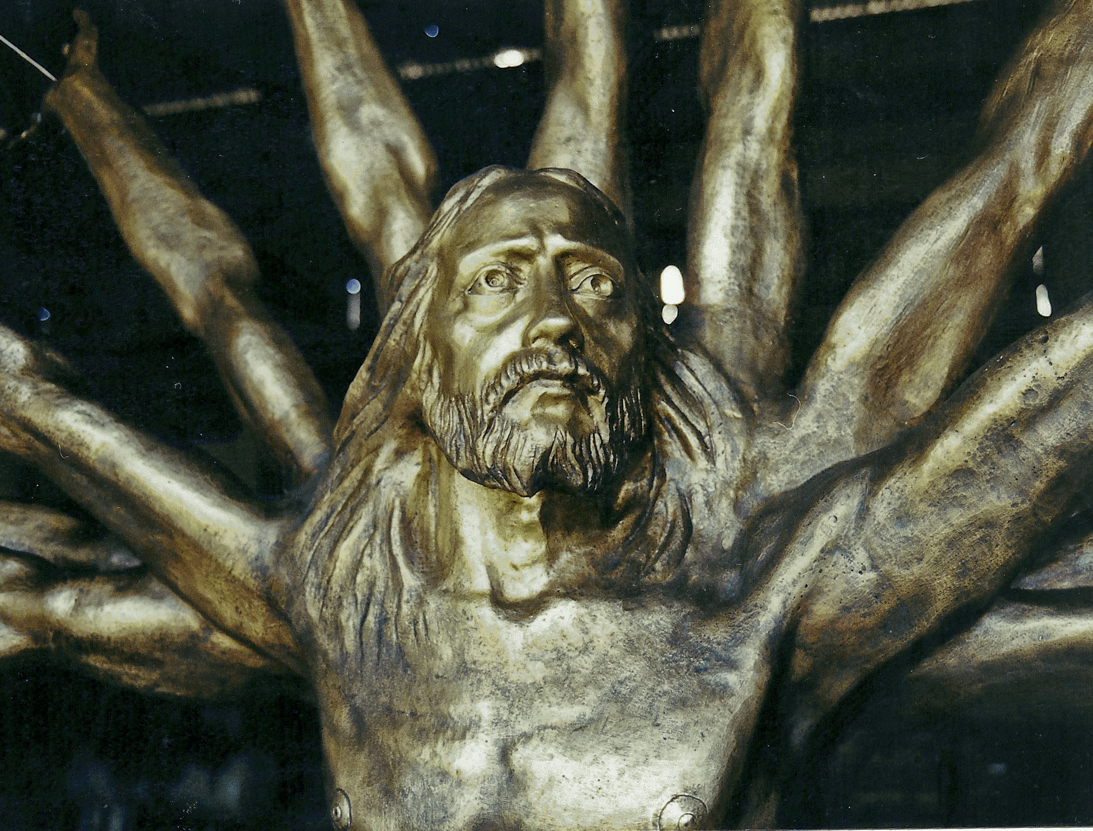 Close-up detail of the golden bronze sculpture's bearded face and multiple radiating arms.