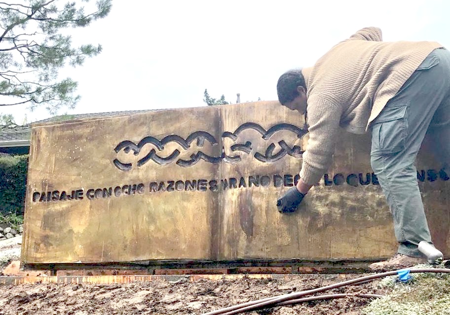 Outdoor photograph of a large bronze relief sculpture shaped like an open book, its Spanish title 'PAISAJE CON OCHO RAZONES PARA NO DECIR LO QUE PIENSO' carved along the base, with a person kneeling beside it applying patina.