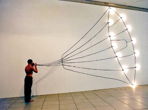 Photograph of a shirtless man in a gallery holding a camera up to his eye, a fan of taut black strings stretching from the camera to a row of bare light bulbs mounted on the wall.