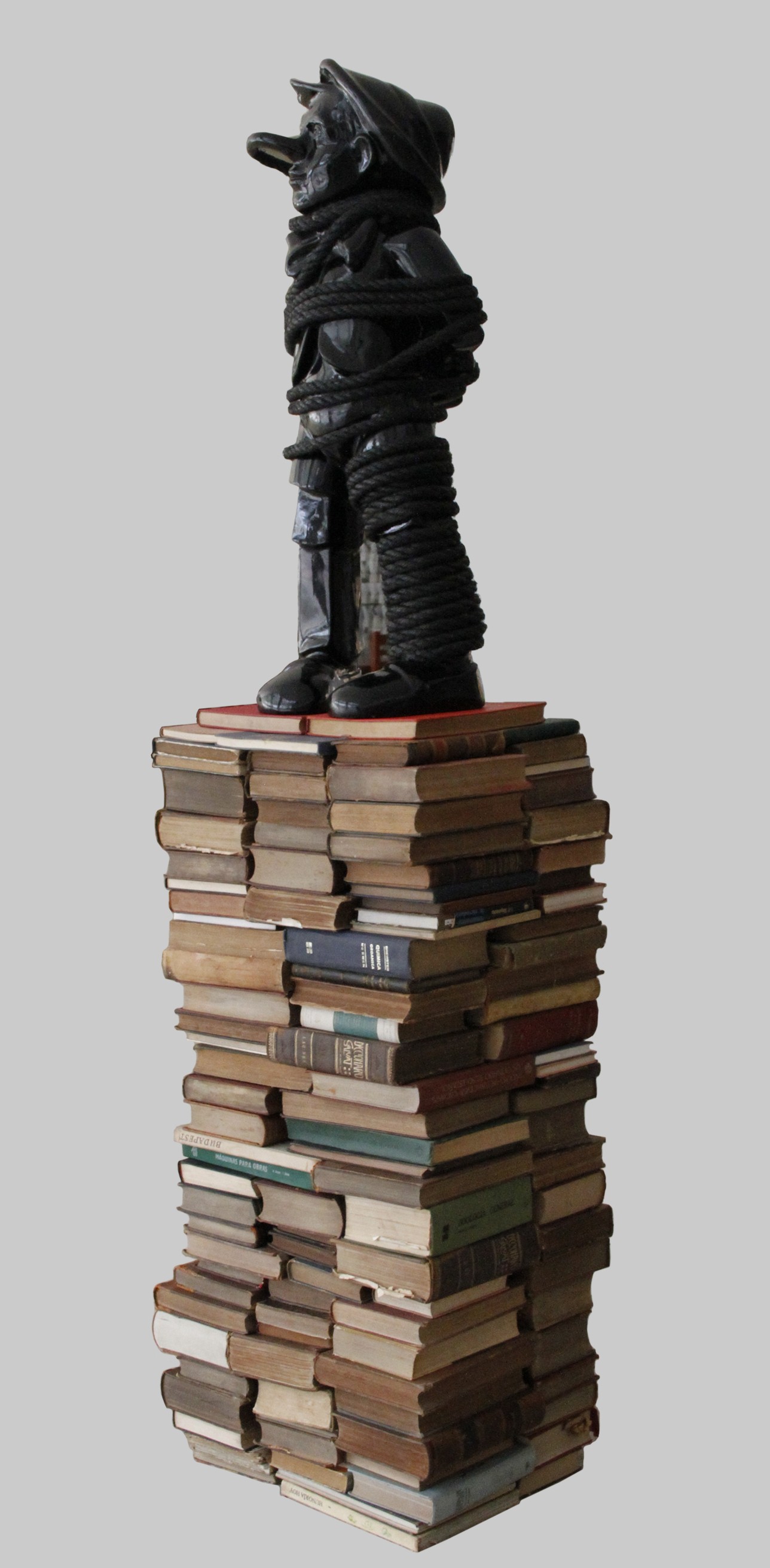 A black Pinocchio-like figure bound in rope, standing atop a tall stack of old books.