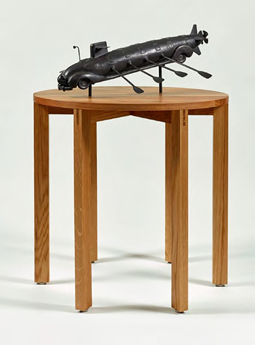 A small bronze maquette of the car-submarine hybrid with oars, displayed on a round wooden table.