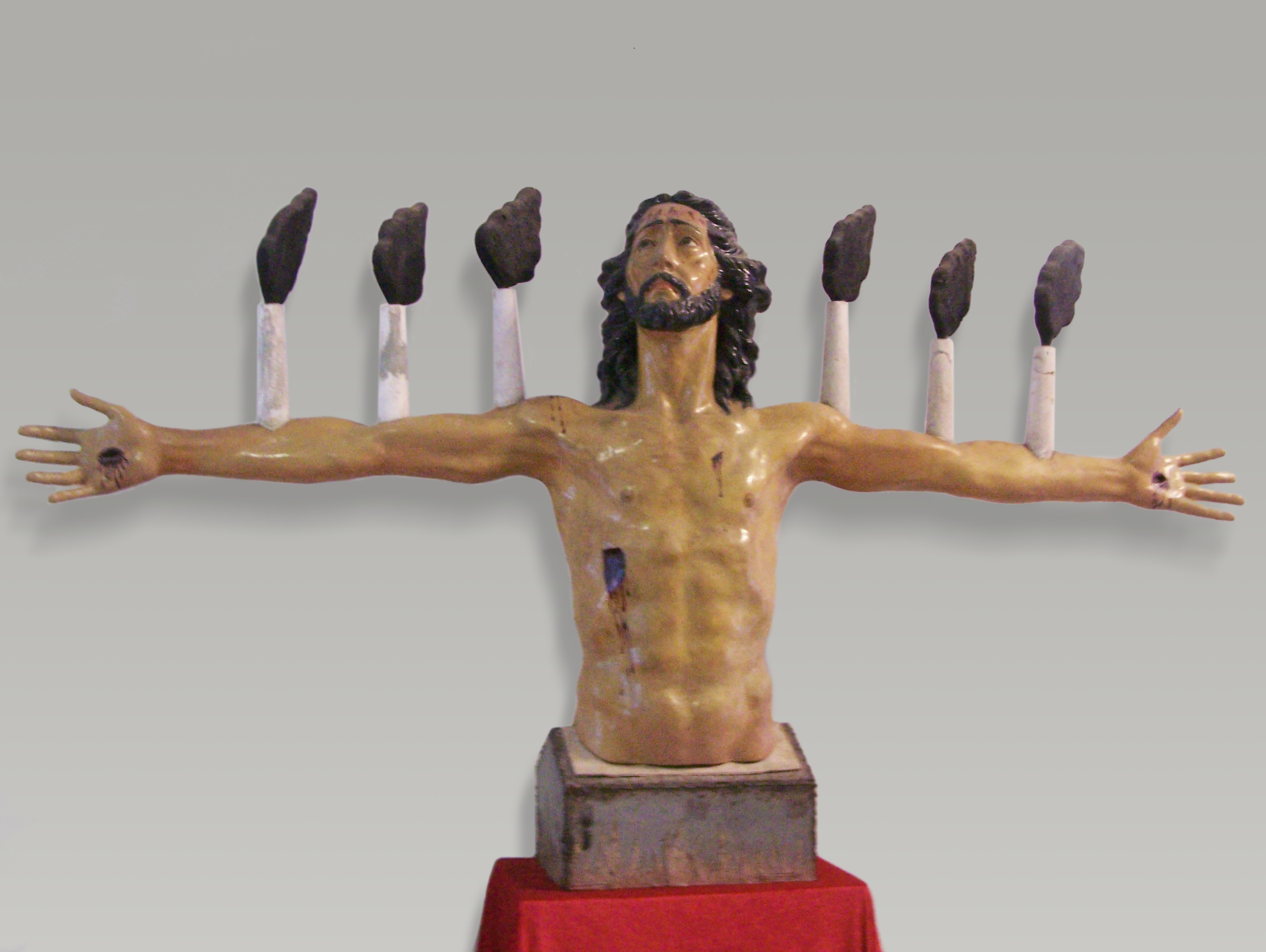 Sculpture of a bare-chested bearded man's torso with arms outstretched, six lit candles rising behind each arm, bleeding wounds on his hands, side, and forehead in the style of a crucified Christ.