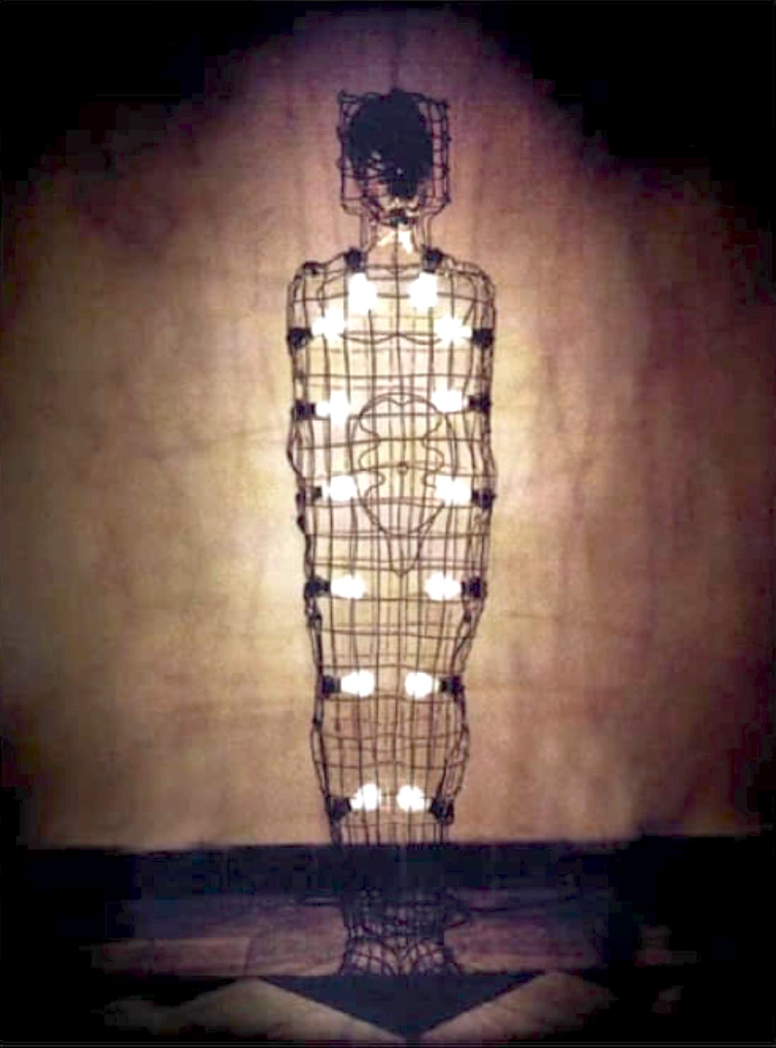 A life-size wire-mesh human figure lit from within, with rows of bare light bulbs glowing through the torso in a cross pattern and a faint heart shape traced on the chest.